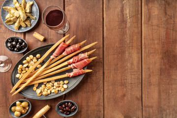 Italian antipasti. Grissini breadsticks with parma ham and roasted almonds, with olives and artichokes, shot from the top on a dark rustic wooden background with wine glasses and a place for text