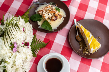 Still life with 2 cakes, cup of coffee and white small flower bouquet on square red-white tablecloth