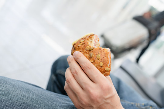 Middle-aged man sitting at airport holding typical German whole grain bun with cheese and ham in his hand and bite taken, a healthy, light and nutritious lunch sandwich or snack to eat while traveling