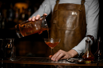 Male bartender pouring an alcoholic drink from the measuring cup to the cocktail glass