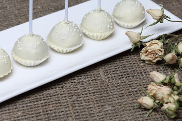 Cake pops in white chocolate glaze. Stand vertically on a stand. Decorated with white decorative sprinkles. Near dry beige rose. On a background of coarse linen fabric.
