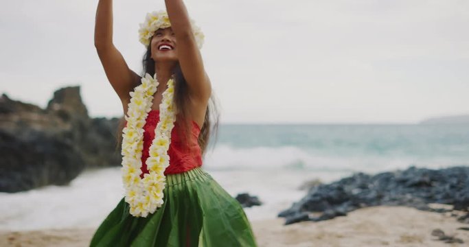 Beautiful smiling woman dancing Hawaiian Hula 'Auana on the beach in slow motion wearing a ti leaf skirt, plumeria lei, and haku