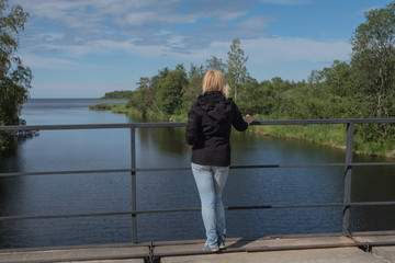 A girl stands on a bridge and looks into the distance. The river flows into the lake. Spring. Sunny day.