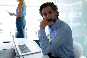 Businessman with hand on chin looking at camera while businesswoman standing in background in office