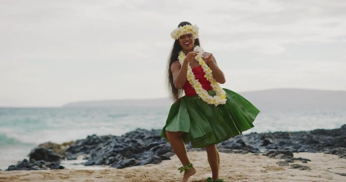 Beautiful smiling woman dancing Hawaiian Hula 'Auana on the beach in slow motion wearing a ti leaf skirt, plumeria lei, and haku