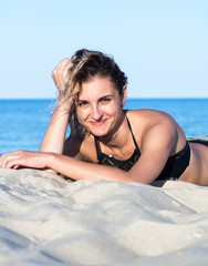 tanned young attractive woman with curly hair in black swimsuit on the beach, lying on the sand