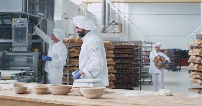 Dancing very attractive baker man with beard while forming pieces of dough for baking bread beside him other old baker preparing the baking molds background workers moving the baked bread