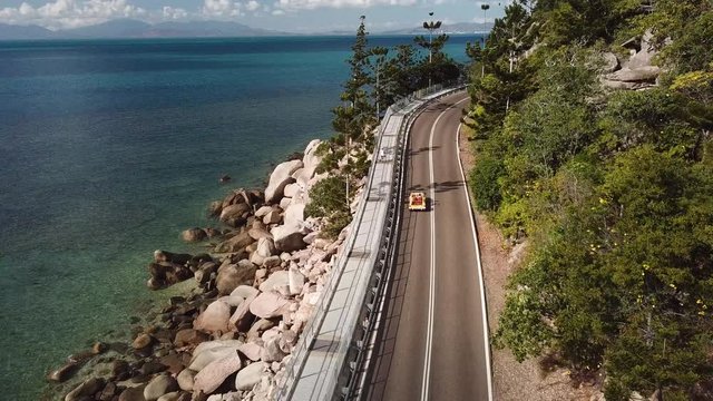Aerial view of open top convertible classic beach buggy driving along coastal clifftop road next to crystal clear tropical water and lush green forest