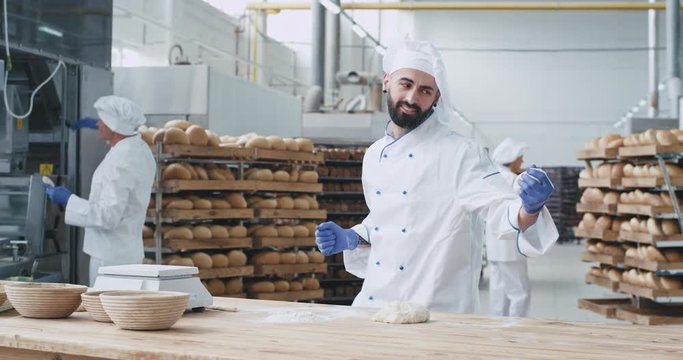 Attractive and charismatic baker very stylish dancing funny in a bakery factory while forming pieces of dough for baking bread, enjoying the time at his work place beside other baker setting the
