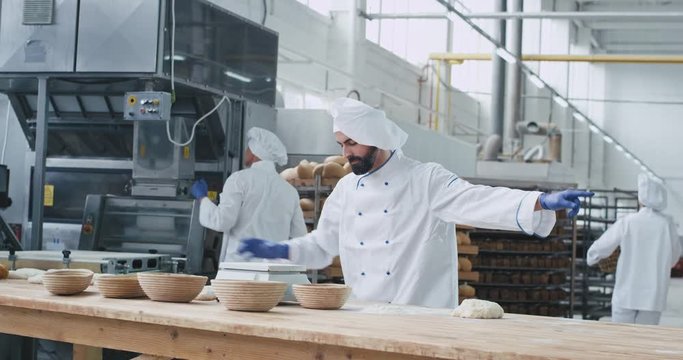 In a bakery industry happy dancing baker have a fun time while preparing the dough for making a bread some workers working concentrated behind him , food industry factory