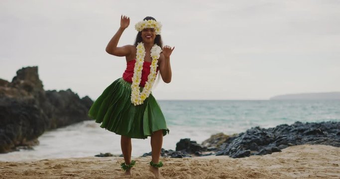Beautiful smiling woman dancing Hawaiian Hula 'Auana on the beach in slow motion wearing a ti leaf skirt, plumeria lei, and haku