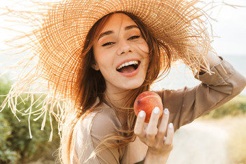 Positive happy cheery woman in hat posing outdoors at the nature green grass near beach eat peach.