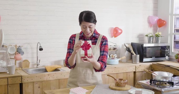 beautiful young asian housewife in apron recieved surprised present while making handmade chocolate. Woman in home kitchen open gift box with cheerful face smiling on valentines day card on table.