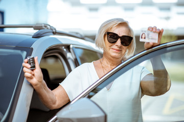 Portrait of a happy senior woman showing driver's license and keys, standing near the car outdoors. Concept of an active people during retirement age