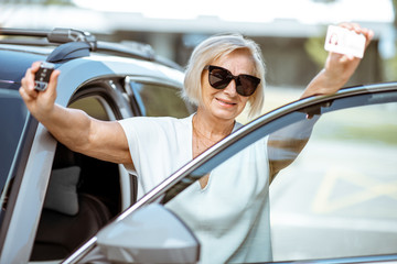 Portrait of a happy senior woman showing driver's license and keys, standing near the car outdoors....