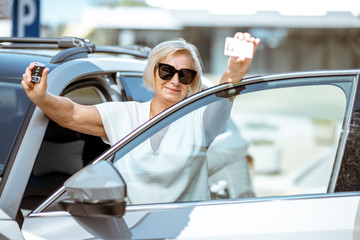 Portrait of a happy senior woman showing driver's license and keys, standing near the car outdoors. Concept of an active people during retirement age