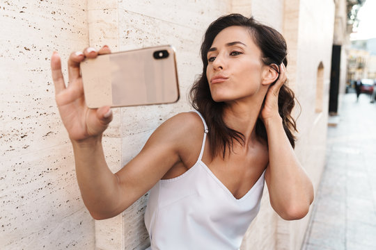 Portrait of alluring woman making kiss face and taking selfie photo on smartphone while standing over wall