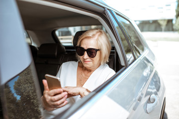 Senior business woman using smart phone while sitting on the back seat of the modern car. Concept of a business life on retirement