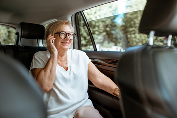 Senior business woman talking with phone while sitting on the back seat in the modern car. Concept of a business life on retirement - obrazy, fototapety, plakaty