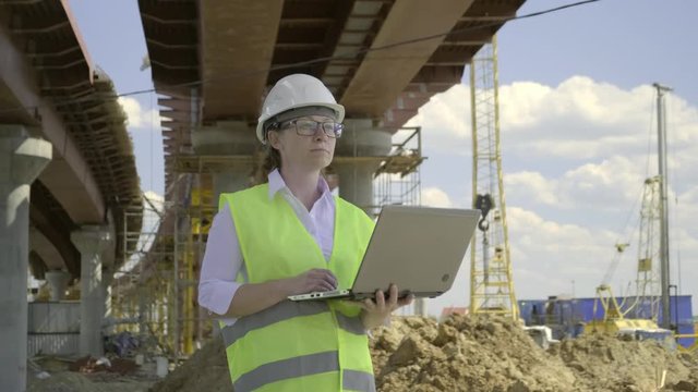 Female foreman working on laptop at bridge construction site. Girl builder controls erection of car road.