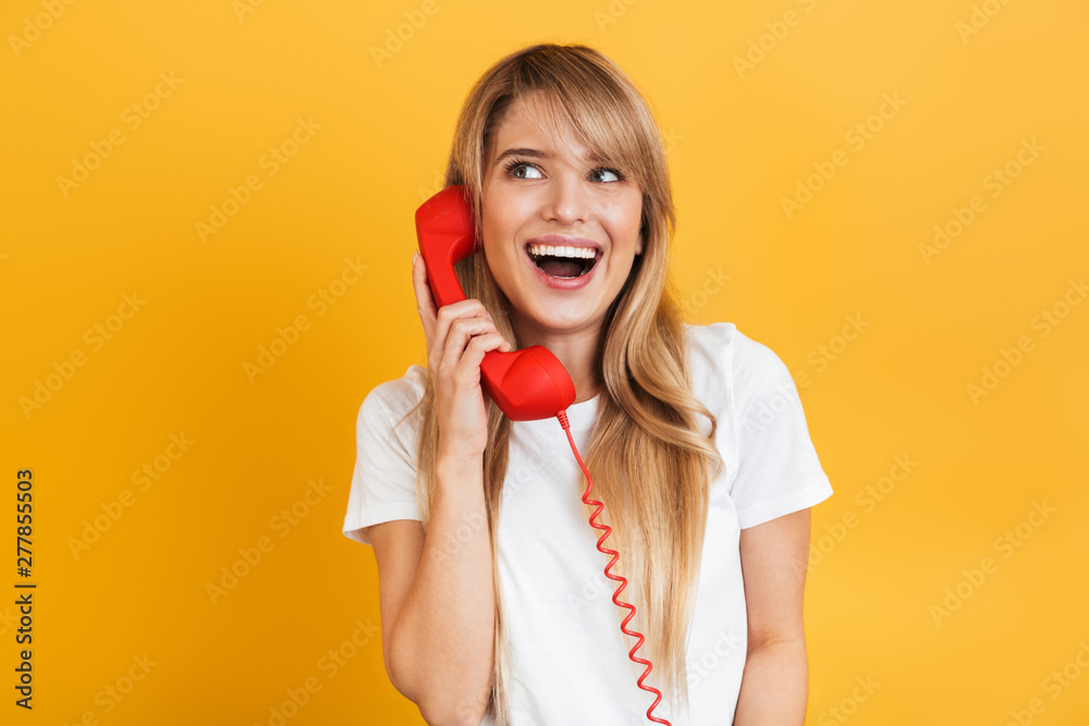 Canvas Prints Smiling emotional happy young blonde woman posing isolated over yellow wall background dressed in white casual t-shirt talking by telephone.