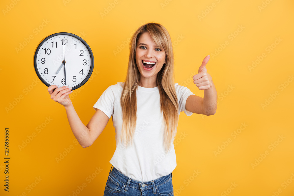 Sticker Happy young blonde woman posing isolated over yellow wall background dressed in white casual t-shirt holding clock showing thumbs up.