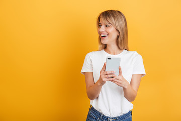 Happy emotional young blonde woman posing isolated over yellow wall background dressed in white casual t-shirt using mobile phone.