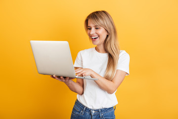 Positive optimistic emotional young blonde woman posing isolated over yellow wall background dressed in white casual t-shirt using laptop computer.