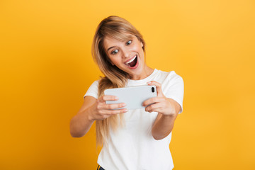 Happy emotional young blonde woman posing isolated over yellow wall background dressed in white casual t-shirt using mobile phone play games.