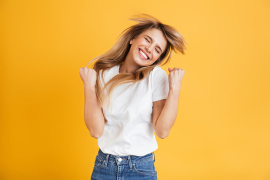 Emotional Young Blonde Woman Posing Isolated Over Yellow Wall Background Dressed In White Casual T-shirt Showing Winner Gesture.