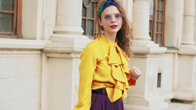 Street fashion portrait of young beautiful elegant lady wearing trendy purple color sunglasses, headband, stylish yellow blouse, holding backpack. Model turns around herself
