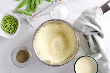 Top view of pot and fresh tasty mashed potatoes in it, bowls with salt and pepper, cup of milk and peeled peas