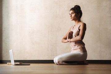 Photo of focused calm woman in sportswear meditating with namaste gesture and using laptop while...