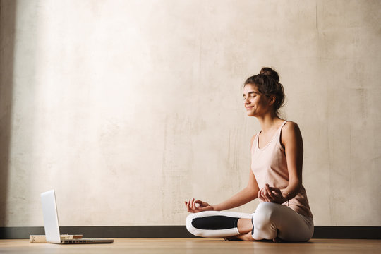 Photo Of Joyful Fitness Woman Meditating With Zen Fingers And Smiling While Sitting On Floor With Laptop At Home