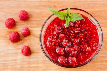 Glass small saucer with raspberry jam with fresh raspberries and mint close-up on wooden background.