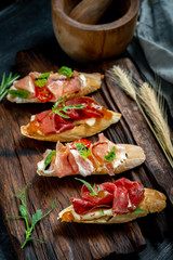 Traditional Bruschetta with Parma dried ham and prosciutto. Italian antipasti set sandwiches with jamon, cottage cheese, arugula, and sun-dried tomatoes on a rustic wooden Board Close up