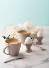 Boiled eggs in ceramic egg cups, two cups of coffee with ceramic spoons on background of beautiful white peony. Breakfast concept. Copy space.