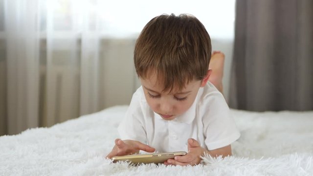 A cute little boy looks into the smartphone screen while lying on the bed. The child plays on the smartphone.