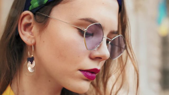 Young fashionable lady wearing trendy purple color sunglasses, colorful headband, posing outdoor, looking at camera. Close up portrait