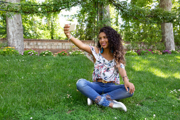 Naklejka na ściany i meble Chica joven latina de piel morena y cabello largo y rizado tomándose un selfie con su smartphone sentada en parque