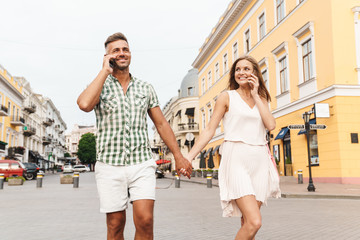 Fototapeta premium Image of happy young couple holding hands together and talking on smartphones while walking through city street