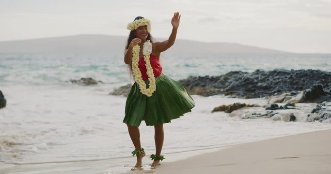 Beautiful smiling woman dancing Hawaiian Hula 'Auana on the beach in slow motion wearing a ti leaf skirt, plumeria lei, and haku