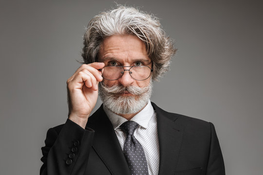 Image of happy adult businessman wearing formal black suit looking at camera and touching his eyeglasses isolated over gray background
