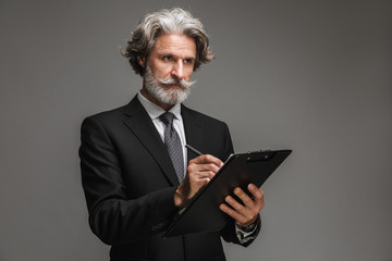 Image of successful adult businessman wearing formal black suit holding clipboard and writing down...