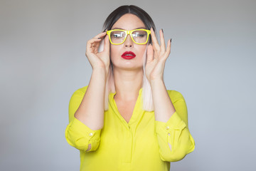 Young beautiful blonde woman in yellow shirt and yellow eyeglasses looking to camera over grey background