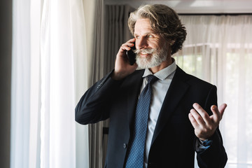 Image of handsome adult businessman wearing black suit talking on smartphone in hotel apartment