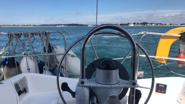 A yacht in light winds, on a sunny day in calm coastal waters. Slowly motoring away from land looking astern showing the autopilot turning the wheel to steer the boat.