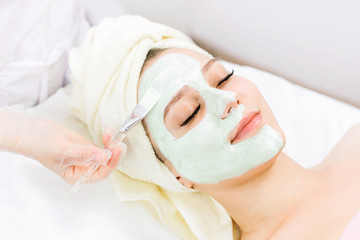 Portrait of beautiful woman laying with towel on the head. Young girl enjoys green cream facial mask. Lady getting spa treatment at beauty salon.