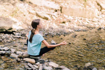 Naklejka na ściany i meble Young girl yoga meditating at sunrise sitting on stones on the shore of a mountain stream. Teenage model meditating in peaceful harmony