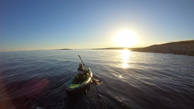 AERIAL: Man paddling his kayak towards the sun setting behind the coastline